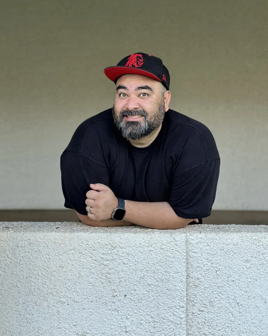 Aaron Uyehara in a black cap, leaning on a concrete wall and smiling